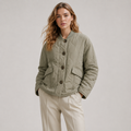 A woman modeling a light olive green quilted jacket with dark buttons, paired with beige trousers, against a neutral light gray background.