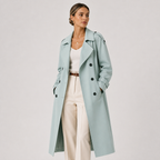 A woman modeling a long, double-breasted trench coat in a pale mint green color, styled with a white top, cream trousers, and a brown belt against a light gray studio background.
