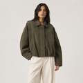 A woman modeling an oversized olive green corduroy jacket with a relaxed fit, paired with cream-colored trousers, against a plain light gray studio background.