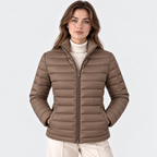 A woman wearing a taupe brown puffer jacket over a cream ribbed turtleneck sweater and light beige trousers, photographed against a clean light gray studio background.