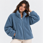A woman wearing a dusty blue sherpa fleece zip-up jacket with a high collar, paired with light beige pants, photographed against a clean white studio background.