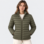 A woman wearing a light olive green puffer jacket over a cream ribbed top and cream pants, photographed against a clean light gray studio background.