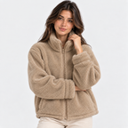A woman wearing a beige sherpa fleece zip-up jacket with a high collar, photographed against a clean light gray studio background.