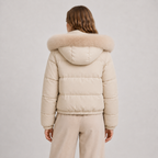 A woman viewed from the back wearing a light beige puffer jacket with a large fluffy beige fur-trimmed hood, paired with matching beige pants, against a light gray studio background.
