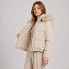 A woman modeling a light beige puffer jacket with a fluffy faux-fur trimmed hood, shown against a neutral light gray studio background.