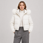 A woman modeling a white puffer jacket with a large fluffy faux-fur trimmed hood, worn over a cream turtleneck and gray trousers, against a light gray studio background.