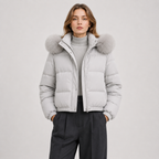 A woman wearing a light gray puffer jacket with a large fluffy faux-fur trimmed hood, layered over a gray turtleneck sweater and dark charcoal trousers, set against a plain light gray studio background.