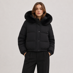 A woman wearing a black puffer jacket with a large black fur-trimmed hood, paired with black trousers, photographed against a plain light gray studio background.