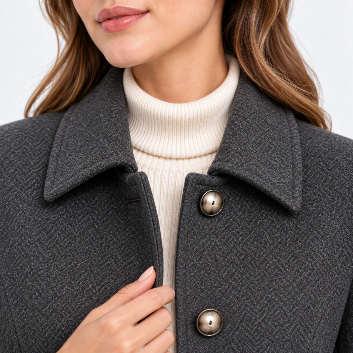 Close-up of a woman wearing a dark gray herringbone wool coat with a wide collar and bronze buttons over a cream ribbed turtleneck sweater.