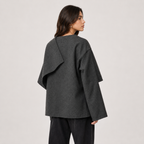 A woman shown from the back wearing a dark charcoal gray draped long-sleeve top and black pants, photographed against a plain light gray studio background.