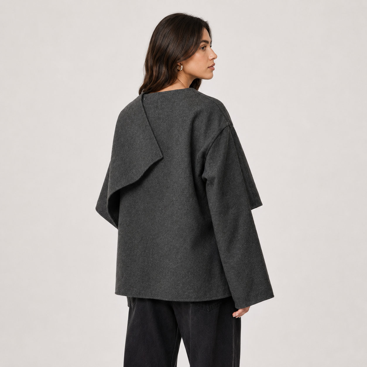 A woman shown from the back wearing a dark charcoal gray draped long-sleeve top and black pants, photographed against a plain light gray studio background.
