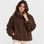 A woman wearing a brown sherpa fleece zip-up jacket with a high collar and side pockets, paired with light beige pants, photographed against a clean white studio background.