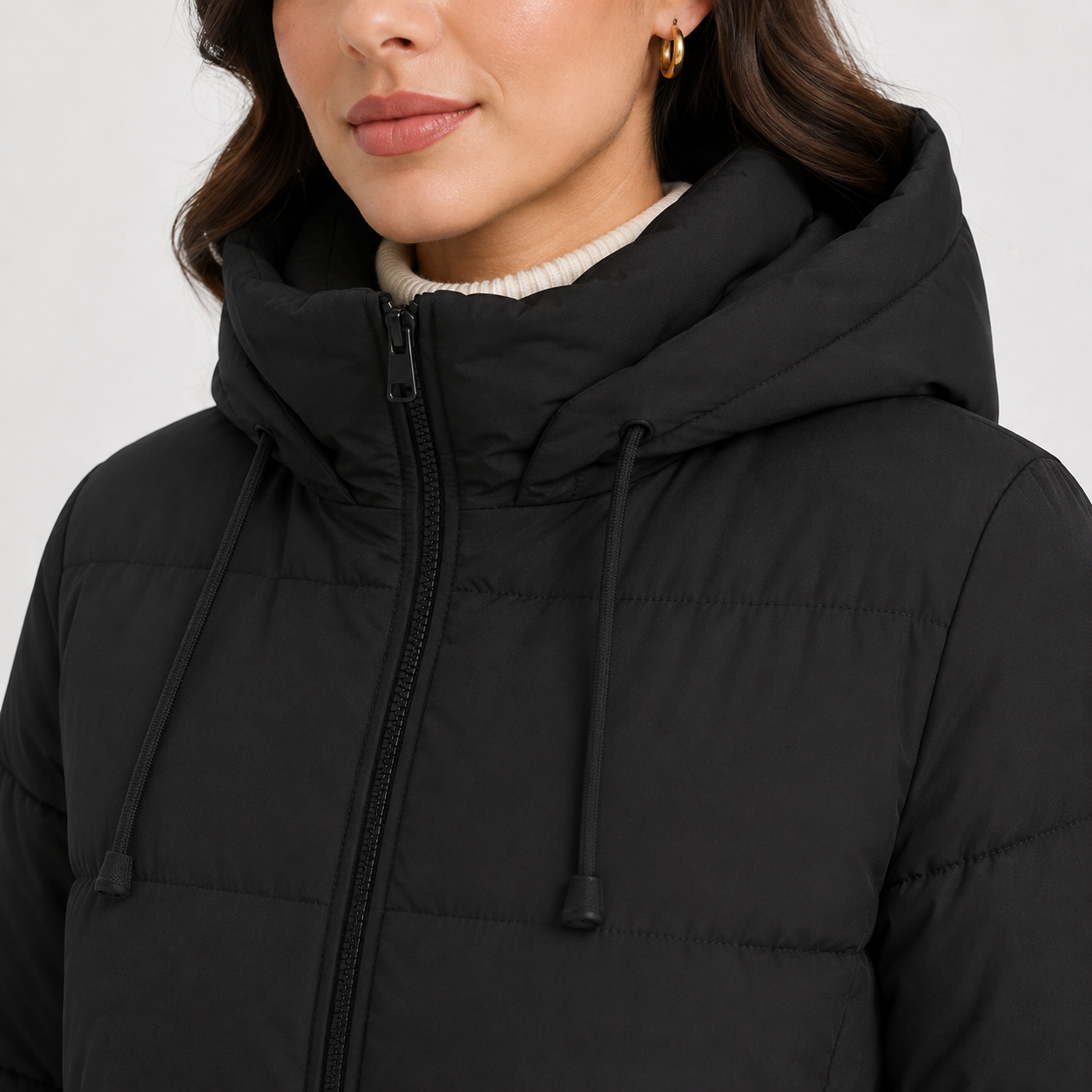 A close-up of a model wearing a black hooded puffer jacket with a front zipper and drawstrings, paired with a cream ribbed turtleneck.