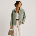 A woman models a light sage green bouclé shearling jacket, styled with a white tank top, cream trousers, a brown belt, and a taupe handbag against a plain light gray studio background.