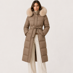 A woman stands against a plain light background wearing a long taupe-brown puffer coat with a large beige faux-fur collar, belted at the waist, paired with cream wide-leg trousers.