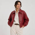 A woman wearing a cropped, shiny burgundy faux-leather jacket over a cream ribbed zip-neck top and cream trousers, standing against a light gray studio background.