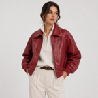 A woman wearing a cropped, shiny burgundy faux-leather jacket over a cream ribbed zip-neck top and cream trousers, standing against a light gray studio background.