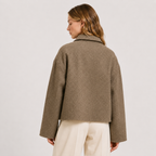 A woman shown from the back wearing a cropped, long-sleeve textured jacket in a warm taupe-brown color, paired with cream-colored trousers, against a plain light gray studio background.