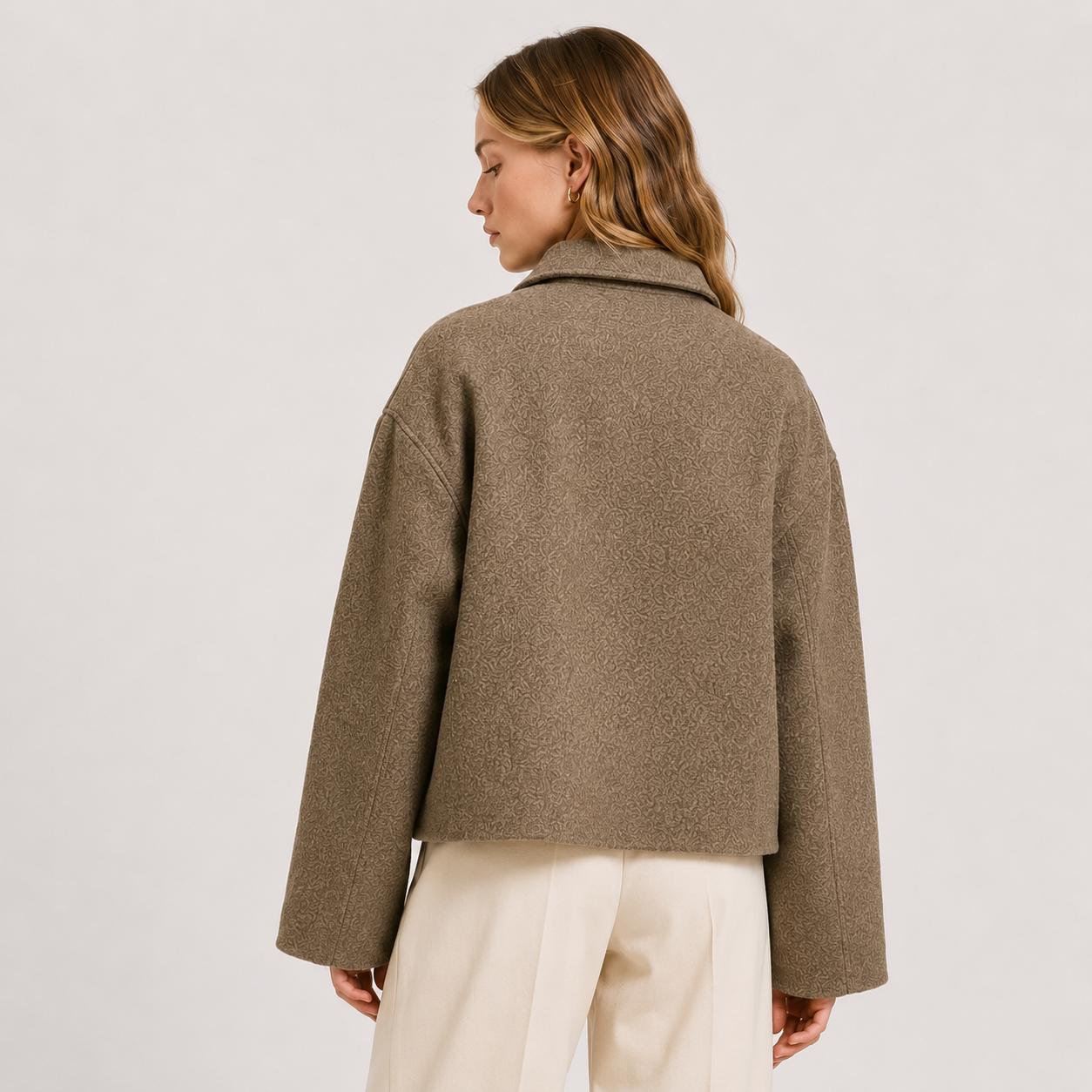 A woman shown from the back wearing a cropped, long-sleeve textured jacket in a warm taupe-brown color, paired with cream-colored trousers, against a plain light gray studio background.