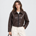 A woman modeling a dark brown faux-leather cropped jacket with a front zipper, collar, and elasticized hem, paired with cream pants against a light gray studio background.