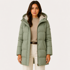 A woman wearing a sage green hooded puffer coat over a beige sweater and cream pants, standing against a plain light background.