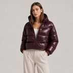 A woman modeling a cropped, shiny dark burgundy puffer jacket with a hood, worn over a white top and cream trousers, against a plain light gray studio background.