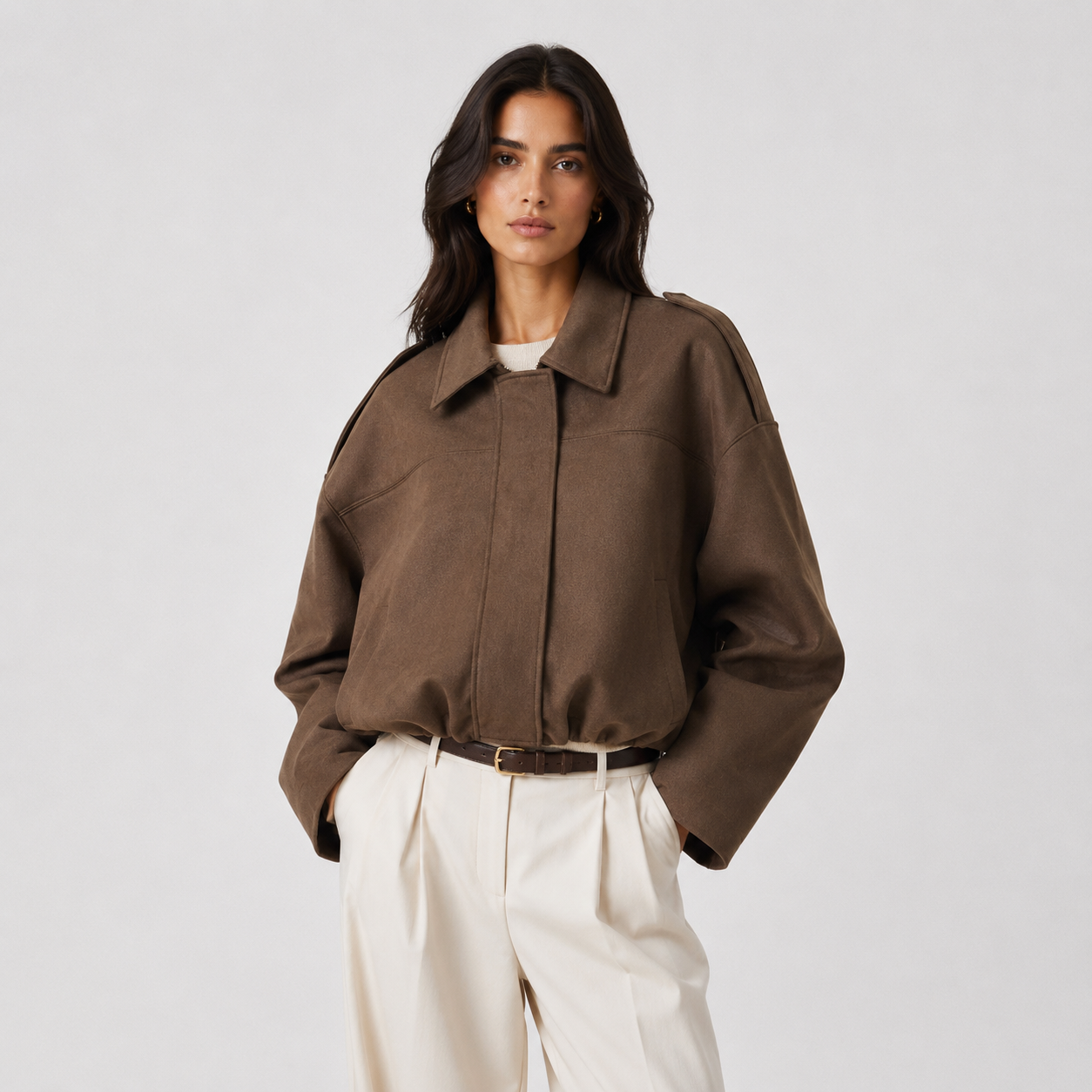 A woman modeling an oversized brown collared jacket with wide sleeves, paired with cream trousers and a thin brown belt, against a light gray studio background.