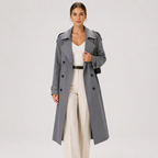A woman modeling a long gray double-breasted trench coat over a white top and cream wide-leg trousers, standing against a light gray studio background.