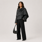 A woman stands against a light gray studio background wearing a dark charcoal draped shawl over a black outfit, holding a black crescent shoulder bag.