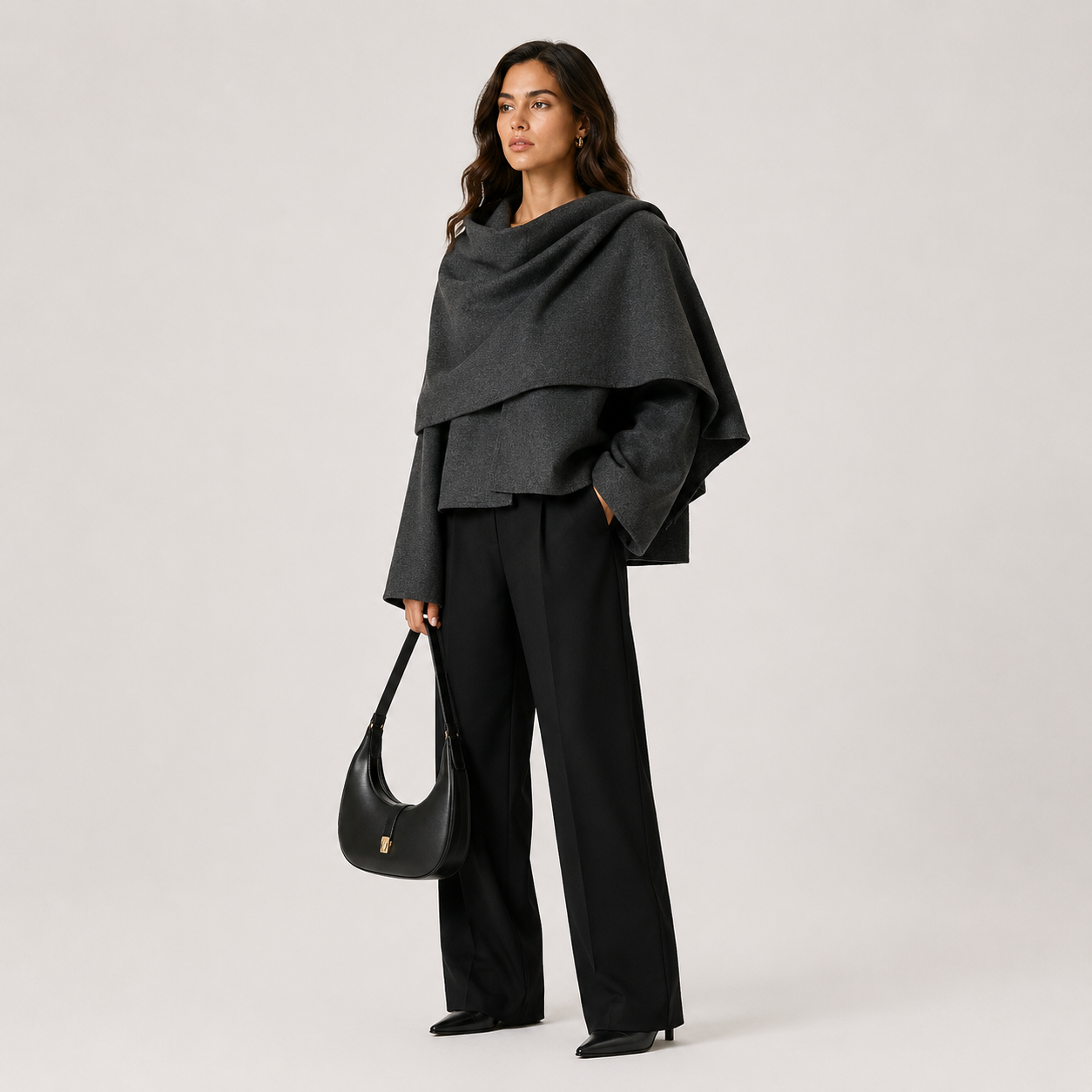 A woman stands against a light gray studio background wearing a dark charcoal draped shawl over a black outfit, holding a black crescent shoulder bag.