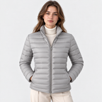 A woman wearing a light gray puffer jacket over a cream turtleneck sweater and beige pants, standing against a plain light gray studio background.