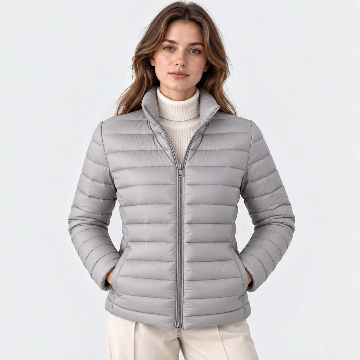 A woman wearing a light gray puffer jacket over a cream turtleneck sweater and beige pants, standing against a plain light gray studio background.
