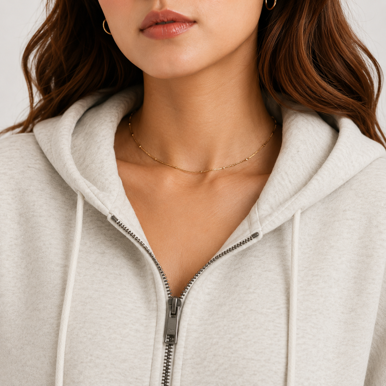 Close-up of a woman wearing a light heather gray zip-up hoodie with white drawstrings and a delicate gold chain necklace.