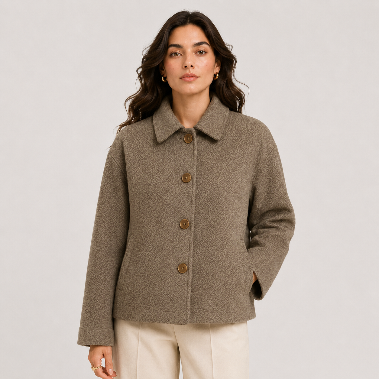 A woman wearing a textured taupe-brown cropped coat with four brown buttons, paired with cream trousers, photographed against a light gray studio background.