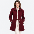 A woman modeling a mid-length burgundy wool-blend coat with silver-tone buttons, worn over a cream turtleneck and cream trousers against a light gray studio background.