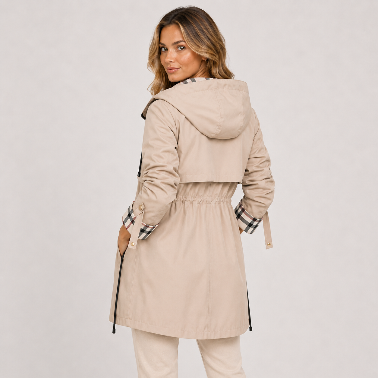 A woman wearing a beige hooded trench coat with a black-and-white plaid lining on the cuffs and collar, shown from the back against a light gray studio background.
