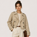 A woman models a beige double-breasted trench jacket over a cream knit top and cream trousers, holding a brown clutch bag against a plain light gray studio background.