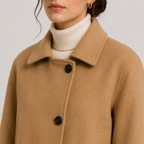 A close-up of a model wearing a camel-colored wool coat over a cream ribbed turtleneck sweater. The coat features a wide collar and two black buttons.