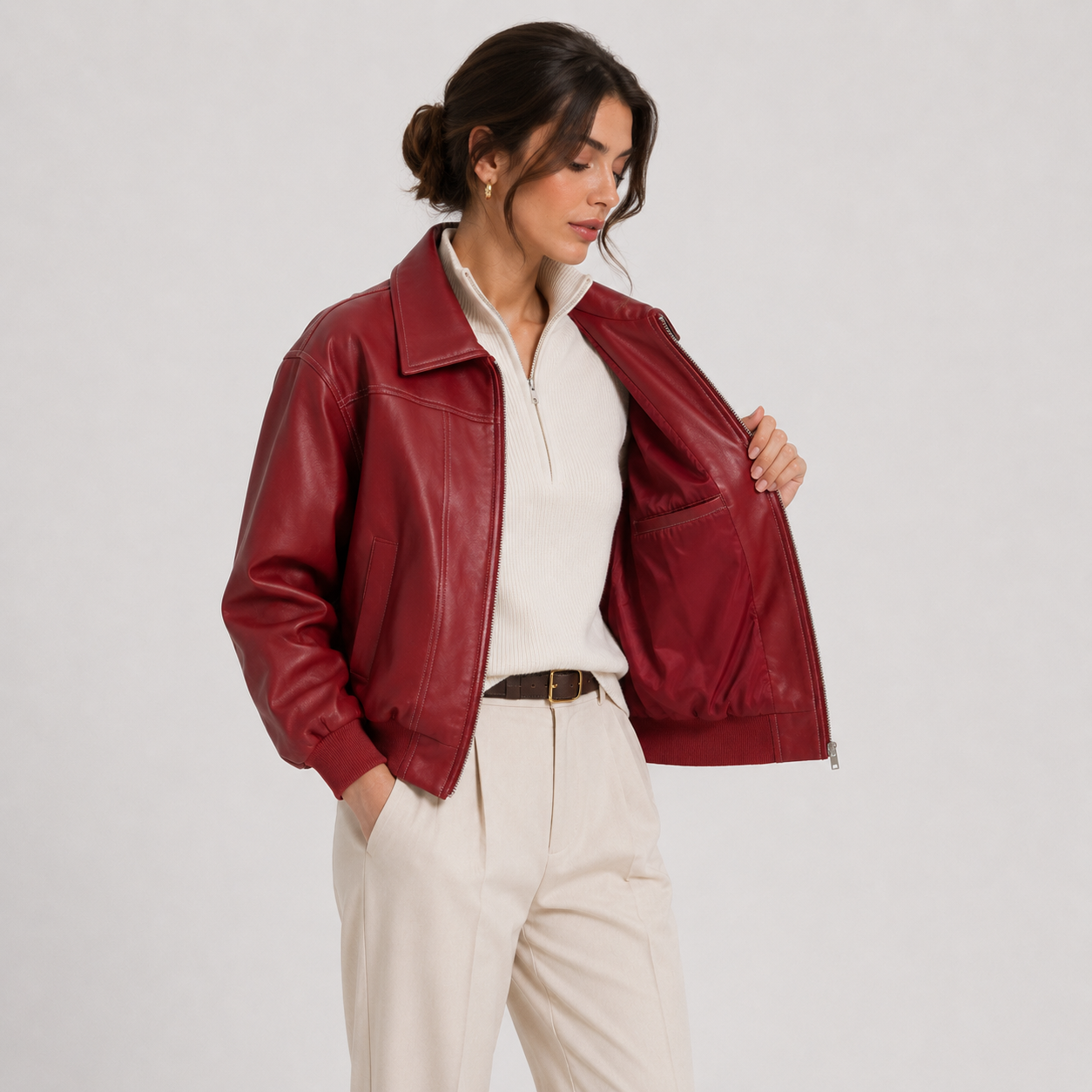 A woman models a cropped, deep burgundy faux-leather jacket with a collar and zippered front, worn open over a white zip-neck top and cream trousers, set against a plain light gray studio background.
