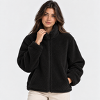 A woman wearing a black sherpa fleece zip-up jacket with a high collar, paired with light beige pants, photographed against a clean white studio background.