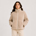 A woman wearing a beige sherpa fleece zip-up jacket with a high collar and front pockets, paired with light beige pants, against a plain light gray studio background.