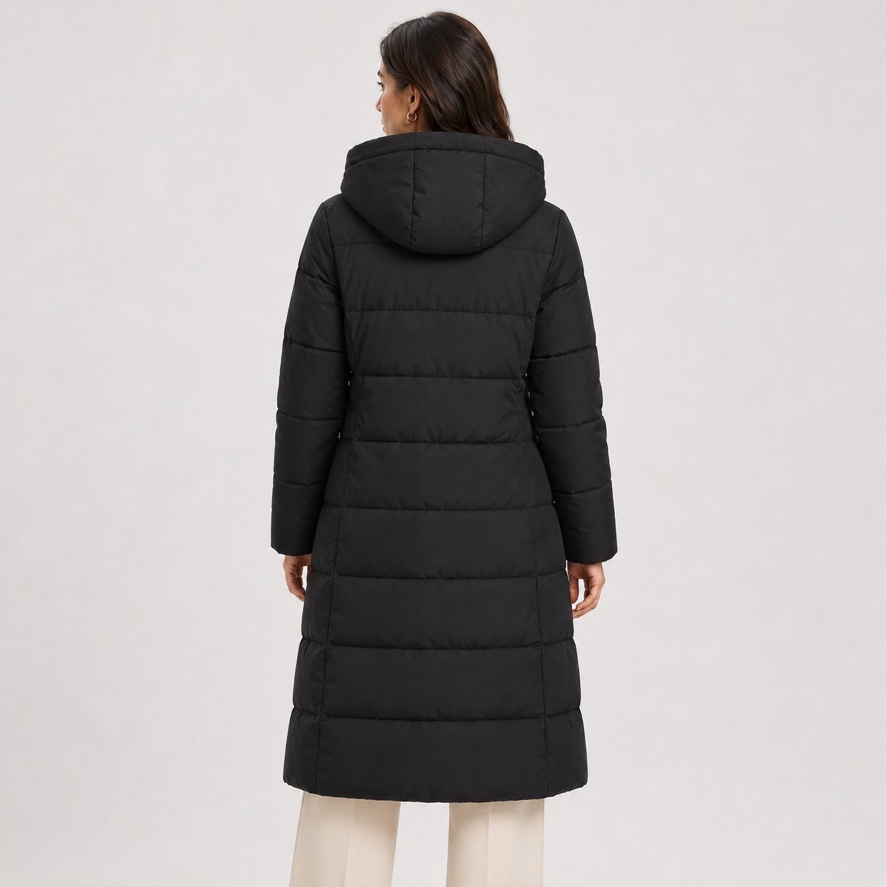 A woman viewed from the back wearing a long black hooded puffer coat over cream pants, photographed against a plain light gray studio background.