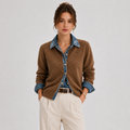 A woman models a brown knit button-up cardigan layered over a blue denim shirt, paired with cream trousers, against a light gray studio background.