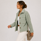 A woman wearing a sage green teddy fleece jacket over a white top and white trousers, holding a taupe shoulder bag against a light gray studio background.