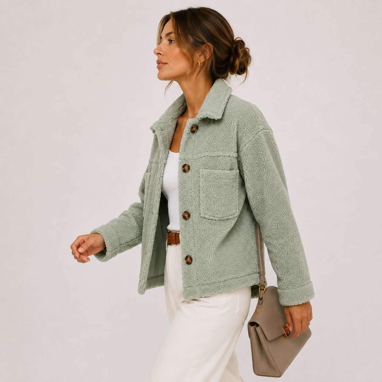 A woman wearing a sage green teddy fleece jacket over a white top and white trousers, holding a taupe shoulder bag against a light gray studio background.