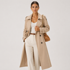 A woman modeling a long beige trench coat over a white top and white trousers, accessorized with a brown belt, gold jewelry, and a brown shoulder bag, against a plain light gray studio background.