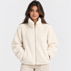 A woman wearing a cream-colored sherpa fleece zip-up jacket with a high stand-up collar and side pockets, paired with beige pants, against a plain white studio background.