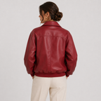A woman shown from the back wearing a glossy burgundy leather jacket and cream-colored trousers, photographed against a plain light gray studio background.