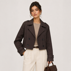A woman models a dark brown cropped double-breasted jacket over a beige top and cream trousers, holding a matching brown handbag against a light gray studio background.