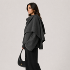A woman modeling a dark gray draped cape-style wrap or poncho over a black outfit, holding a black crescent shoulder bag against a plain light gray studio background.